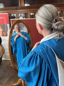 A photo of Professor Karla Pollmann in blue and white academic robes, looking into a mirror before one of Bristol's July 2022 graduation ceremonies. The photo has been taken from behind, and Karla's reflection can be seen in the mirror.