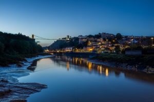 The River Avon at low tide, with the Clifton Suspension Bridge above. It is dark and the lights from nearby buildings are reflected in the water